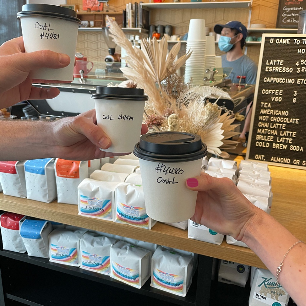 Three hands holding hot oat milk lattes in three white cups with black lids in front of a barista wearing a mask and a variety of retail coffee bags on shelves at Cognoscenti Coffee on Washington in Culver City 
