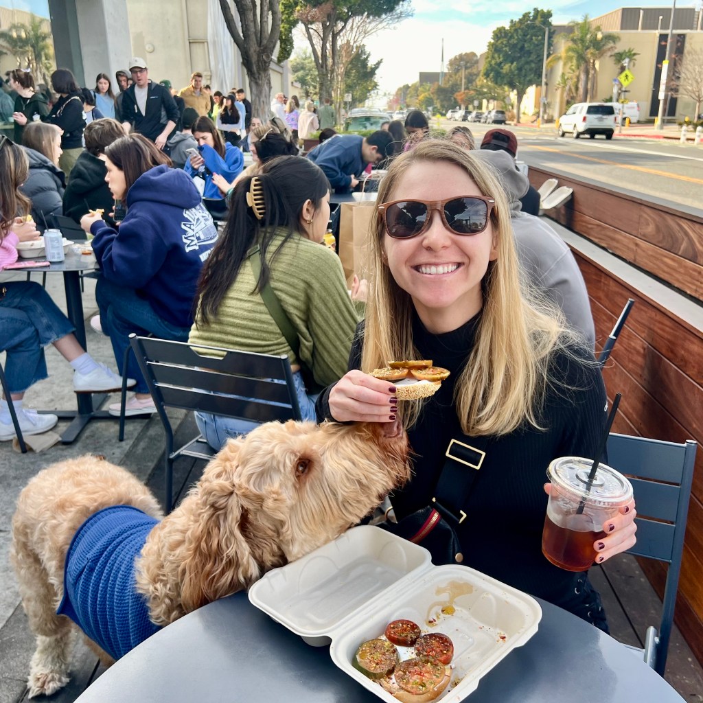 Dog trying to get The Scarlett bagel with heirloom tomato, lemon zest, chili flakes and vegan cream cheese on a gluten free bagel from smiling blonde white girl wearing sunglasses sitting outside at Layla Bagels with the long line in the left side of the photo